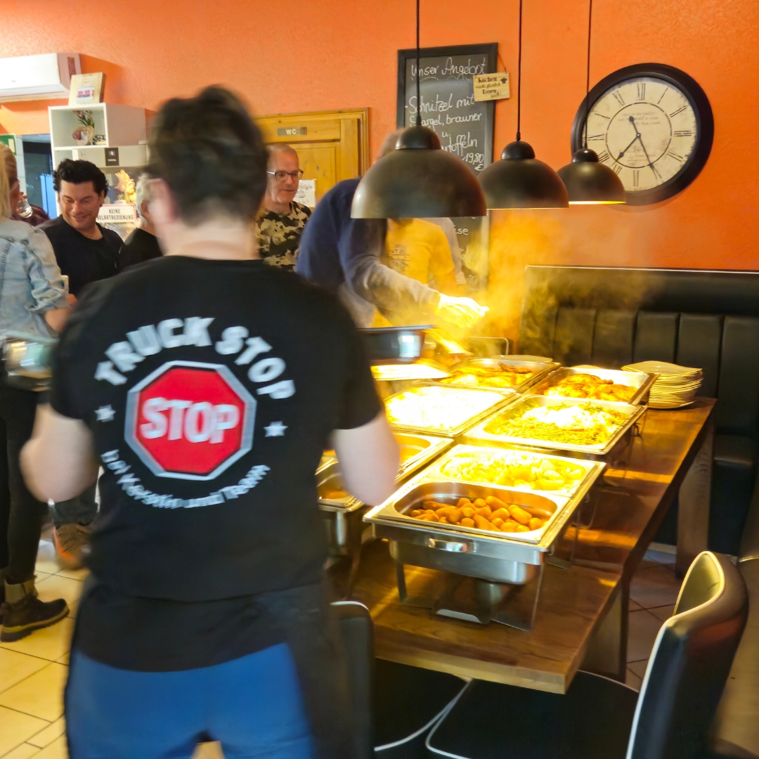 Zu sehen ist ein dampfendes, warmes Buffet, von dem sich mehrere Personen Speisen auf ihre Teller nehmen.