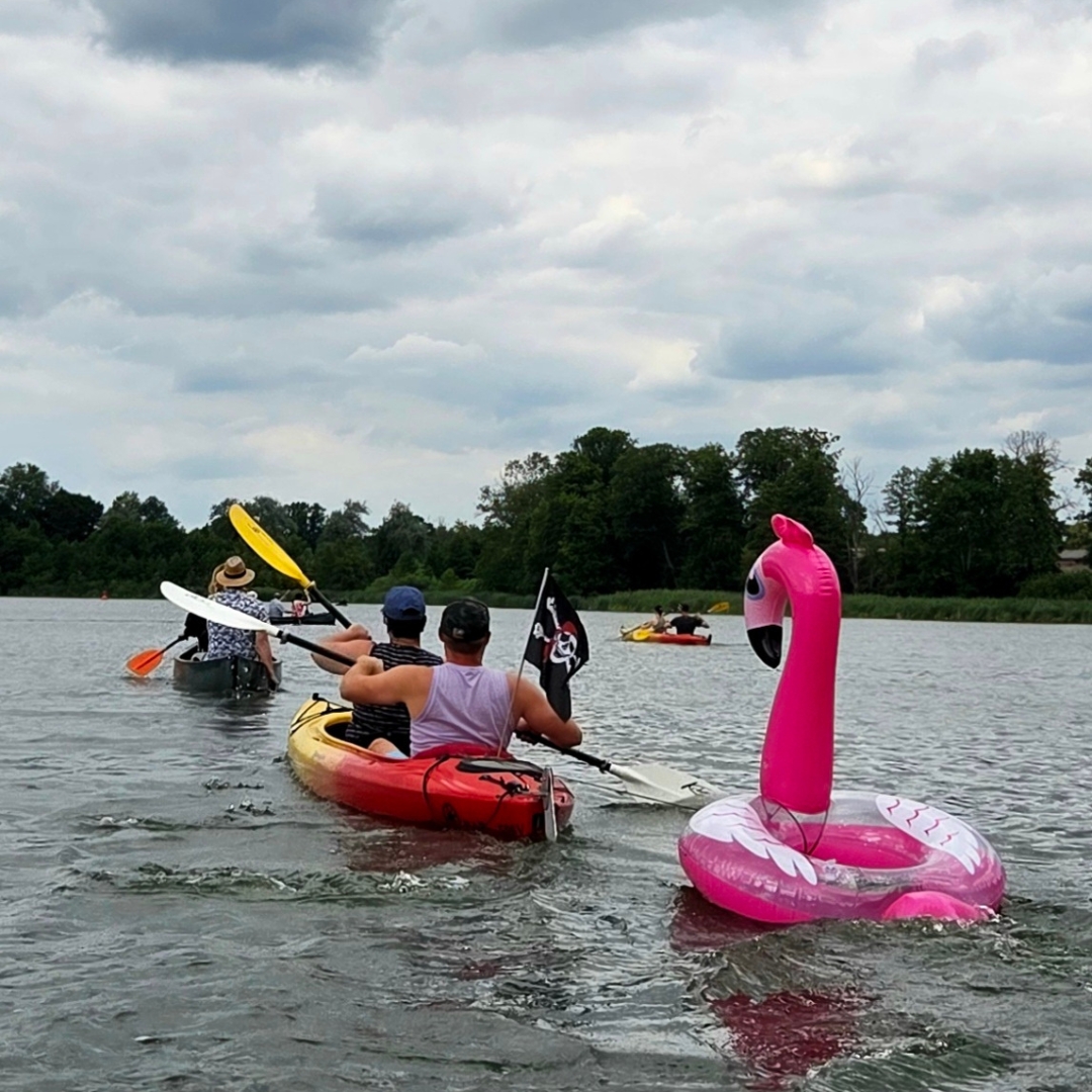 Zu sehen sind mehrere Personen, die mit Kajaks über einen See fahren. Im Wasser ist auch ein  Schwimmreifen in Form eines Flamingos zu erkennen