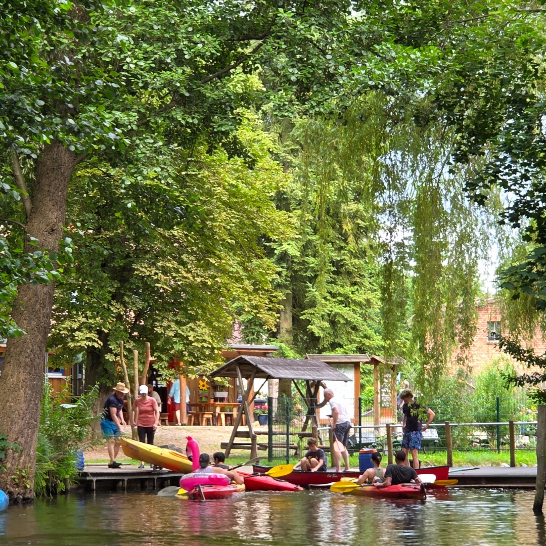 Zu sehen ist ein Park am Wasser. Im See befindet sich eine Person neben einem Schwimmreifen in Form eines Flamingos, während sich weitere Personen am Ufer aufhalten.