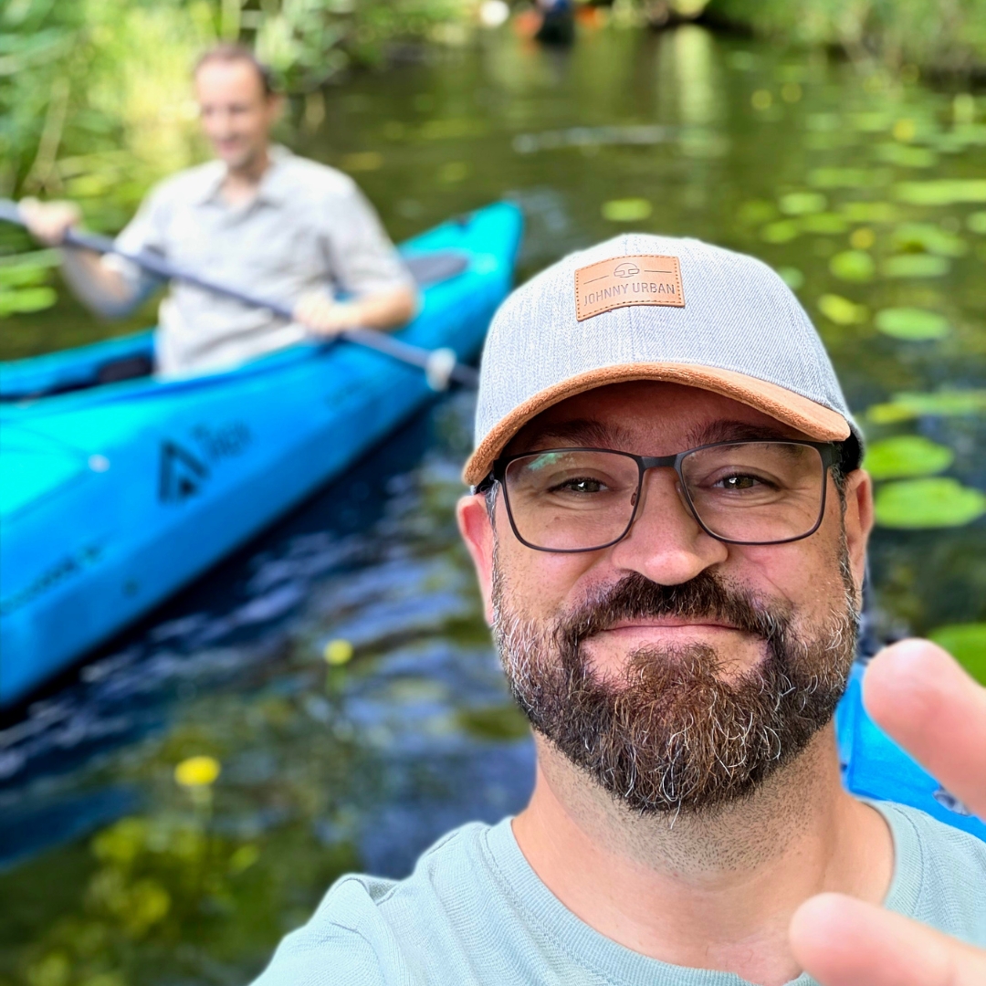 Zu sehen ist eine Person, die lächelnd in die Kamera blickt, im Hintergrund ist eine weitere Person in einem Kajak erkennbar.