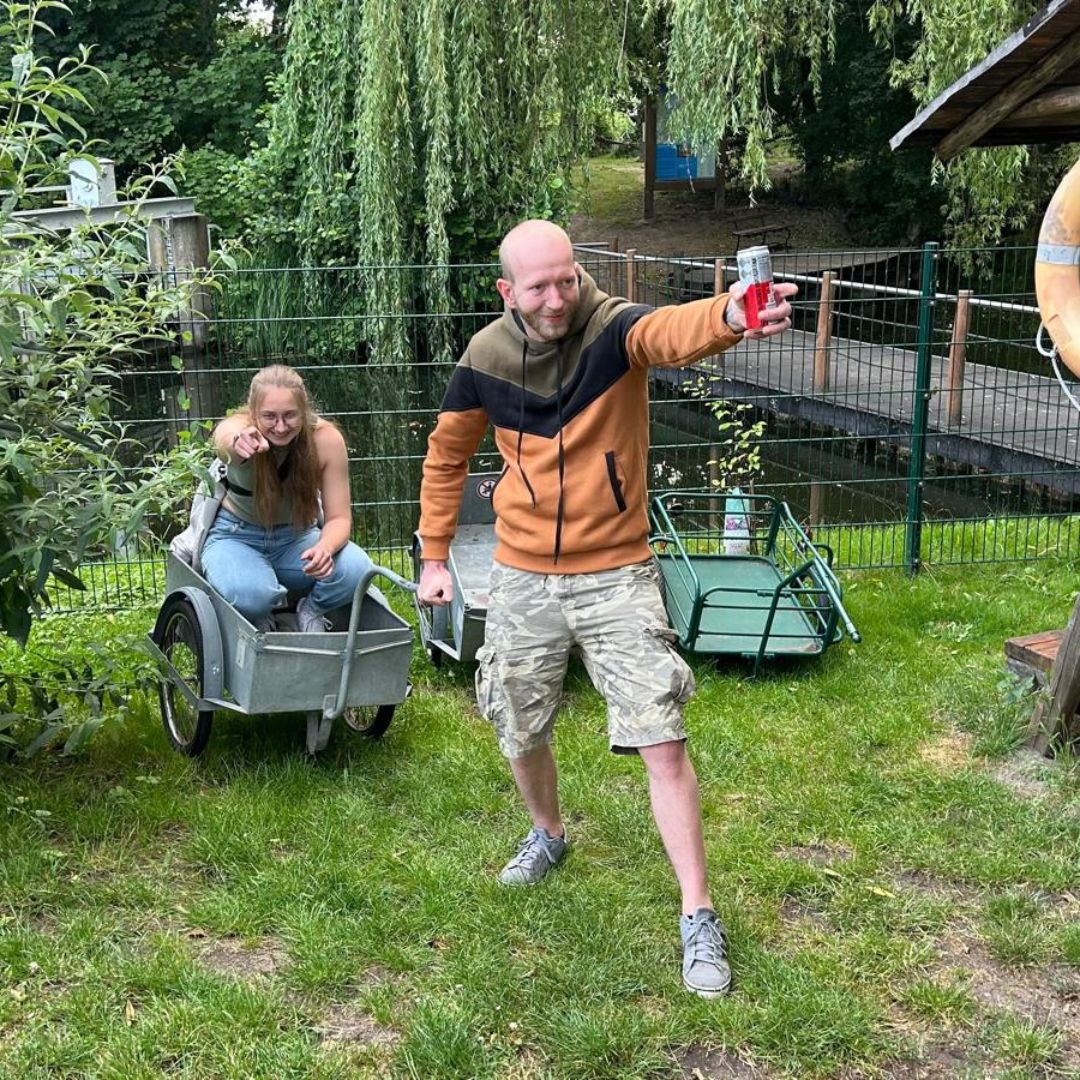 Auf einer Wiese befinden sich zwei Personen: Eine sitzt in einem Anhänger, während die andere diesen festhält und mit erhobenem Arm eine Getränkedose präsentiert.