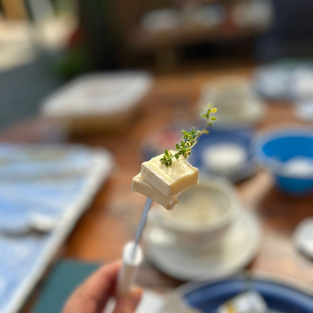 Zu sehen ist ein hochgehaltenes Messer, auf dessen Spitze zwei Käsewürfel aufgespießt sind, im verschwommenen Hintergrund ist ein gedeckter Tisch zu erkennen.