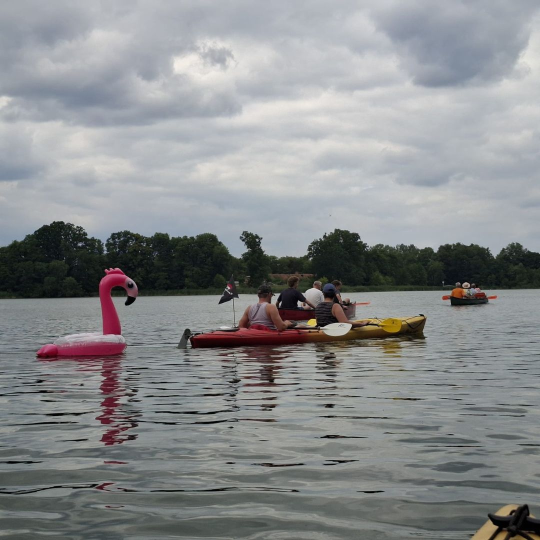 Zu sehen sind mehrere Personen, die mit Kajaks über einen See fahren. Im Wasser ist auch ein  Schwimmreifen in Form eines Flamingos zu erkennen