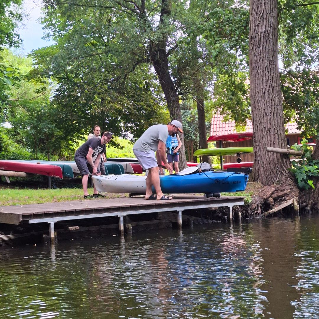 Vier Personen sind zu erkennen, von denen zwei gemeinsam ein Kajak zum Seeufer tragen.