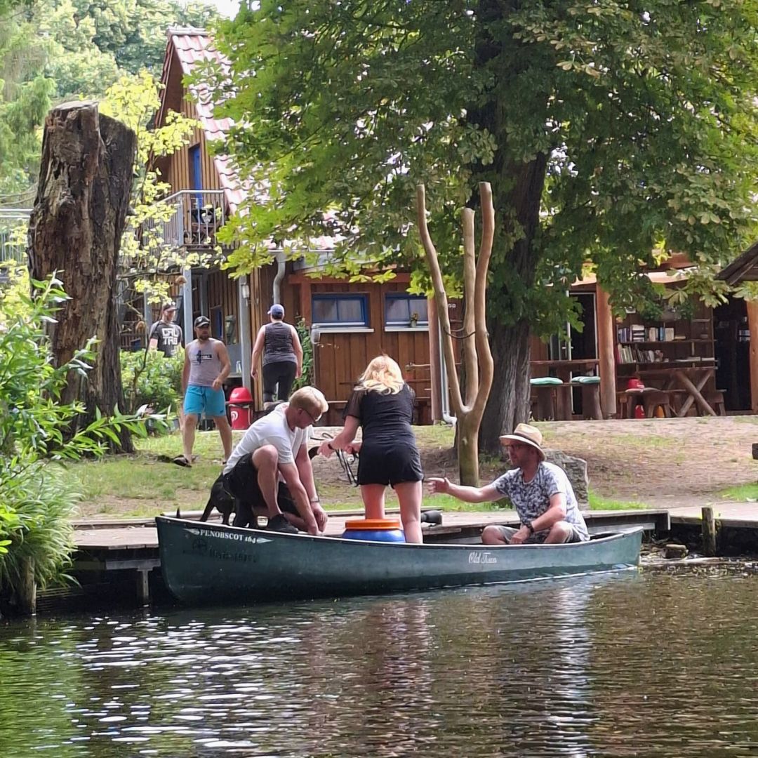 Es sind fünf Personen zu erkennen, von denen drei in einem Kajak auf dem See sind