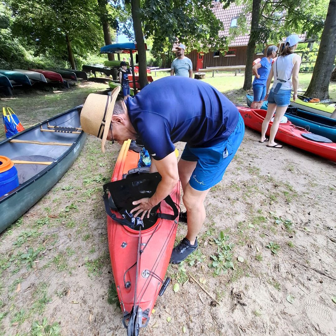 Es sind vier Personen zu erkennen, wobei sich eine von ihnen vor einem Kajak nach vorn beugt.