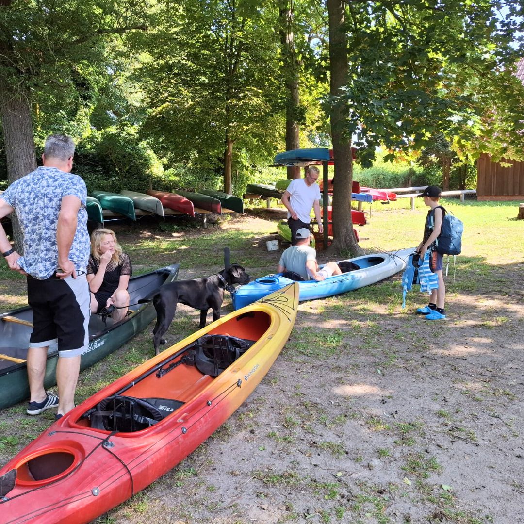 Es sind fünf Personen zu erkennen, die sich in der Nähe von drei auf dem Boden liegenden Kajaks befinden.