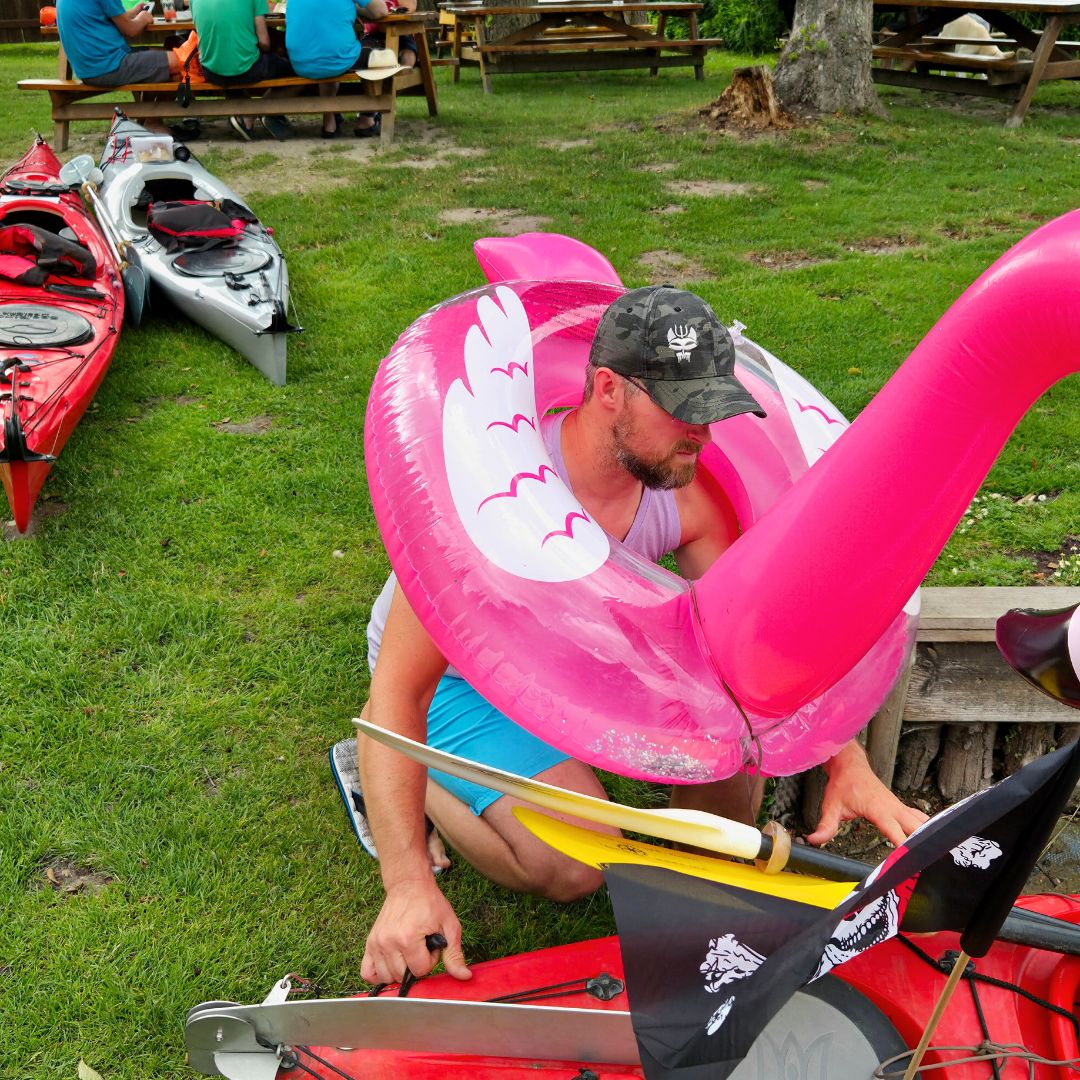 Zu sehen ist eine Person, die auf einer Wiese vor einem Kajak kniet und einen Schwimmreifen in Form eines Flamingos um den Hals trägt.