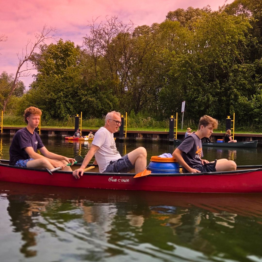 Drei Personen befinden sich in einem Kajak auf dem See, während der Himmel in sanften Rosatönen leuchtet.